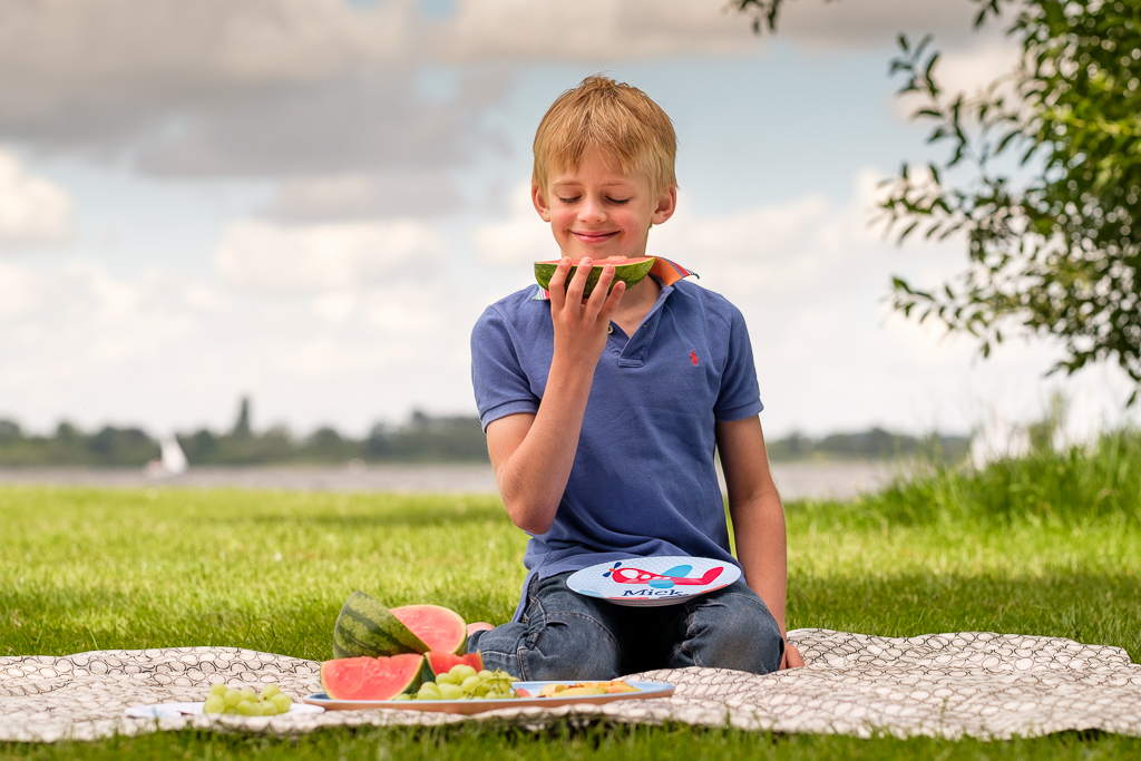 Jongetje wat op picknickkleed zit met bordje van KijkMijne op schoot. Eet een watermeloen. Foto voor de website door Sandra Stokmans Fotografie.