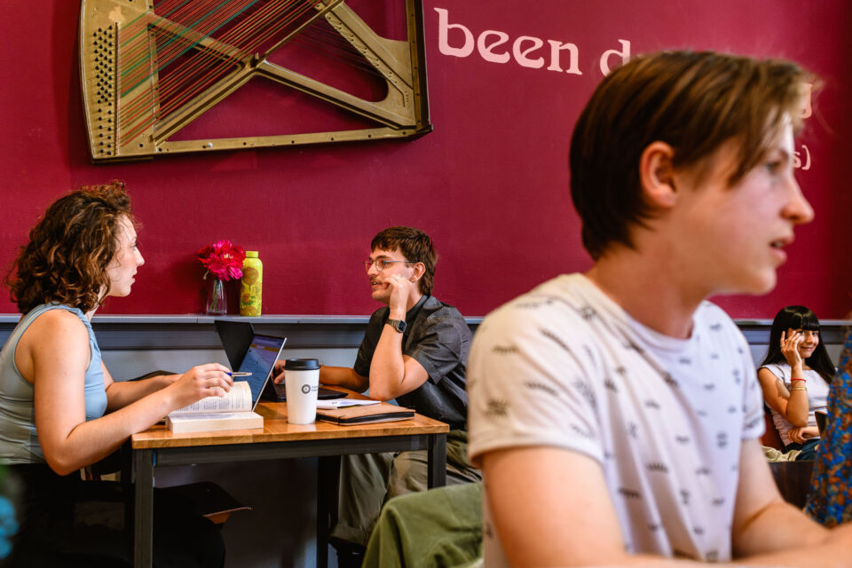 Een typische 'Sandra' moment van aandacht. Voorgrond: een student die aandachtig kijkt, terwijl achter hem twee studenten geconcentreerd aan tafel werken en een meisje gezellig kletst. Deze spontane momentopname vangt de levendige sfeer in het cultuurcentrum van de universiteit.