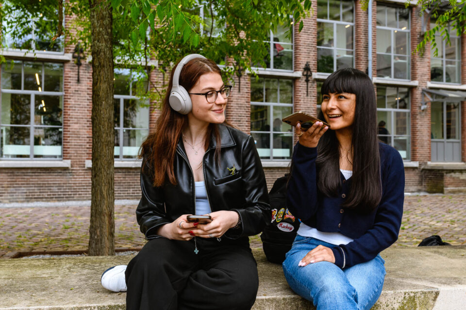 Een ontspannen moment buiten, waar de ene student geniet van muziek terwijl de ander kletst via de telefoon. Dit documentaire beeld legt de balans tussen samenzijn en individuele momenten vast voor de de universiteit beeldbank.