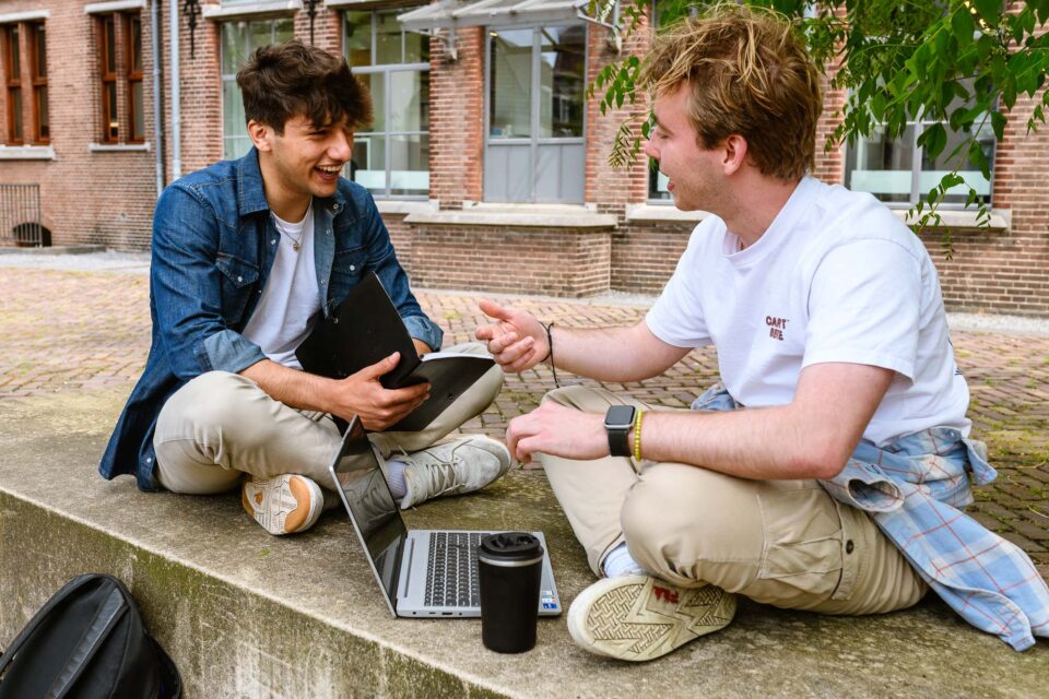Een spontaan moment van twee studenten die zittend op een muurtje aan het werk zijn buiten. Dit beeld benadrukt de veelzijdigheid van studeren aan de Universiteit.