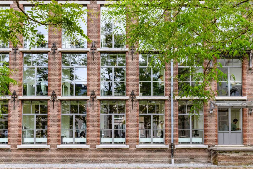 Een portret van het universiteitsgebouw aan het Janskerkhof in Utrecht, met glinsterende ramen die de schittering van de bladeren van de bomen weerspiegelen. Achter het glas zijn verschillende studerende personen zichtbaar, wat de levendige sfeer van de universiteit benadrukt.